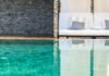 A green tiled swimming pool with a sofa set on the pool coping next to a stone wall.